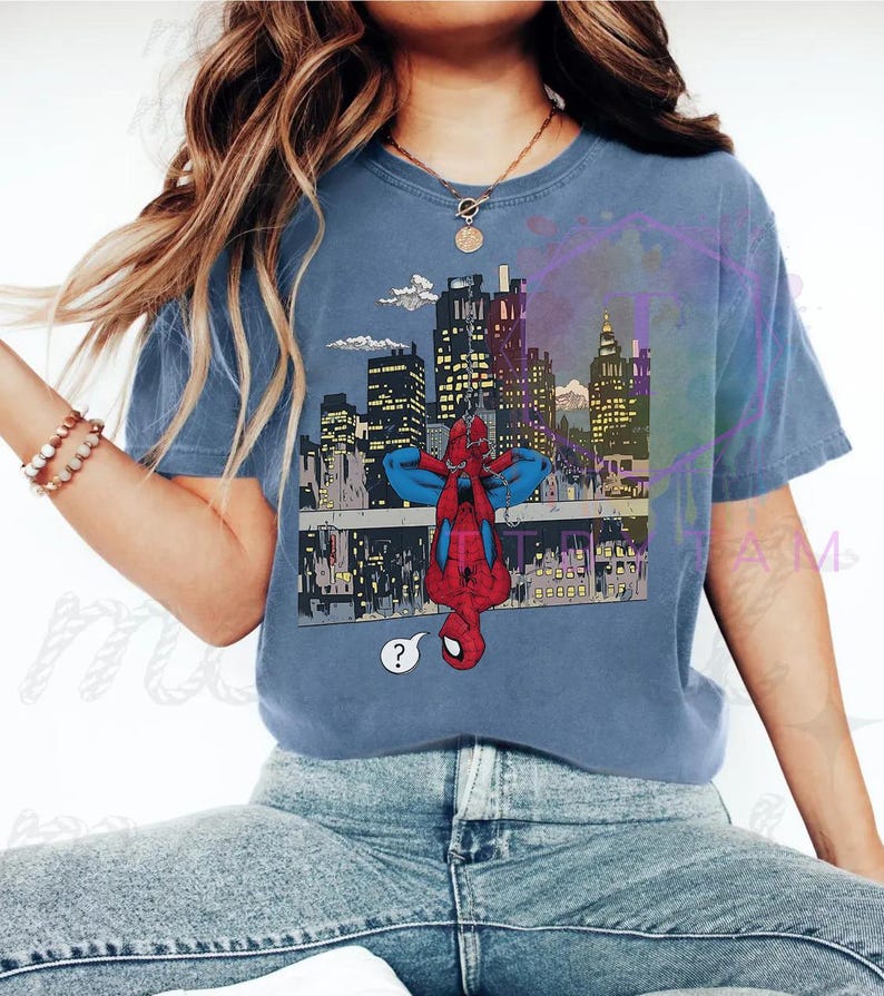 May include: Blue t-shirt featuring a comic-style illustration of Spider-Man hanging upside down in front of a city skyline. The graphic includes a question mark speech bubble. The shirt is a solid blue color.