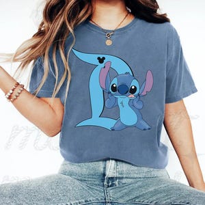 Peut inclure: Un t-shirt bleu &agrave; manches courtes orn&eacute; d'un motif repr&eacute;sentant le personnage Stitch de Lilo et Stitch. Le personnage est plac&eacute; devant une grande lettre D bleu clair qui comporte une petite ic&ocirc;ne noire de Mickey Mouse en haut.