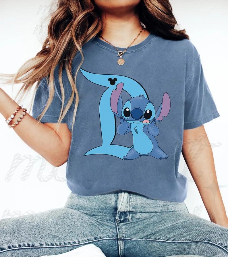 Peut inclure: Un t-shirt bleu &agrave; manches courtes orn&eacute; d'un motif repr&eacute;sentant le personnage Stitch de Lilo et Stitch. Le personnage est plac&eacute; devant une grande lettre D bleu clair qui comporte une petite ic&ocirc;ne noire de Mickey Mouse en haut.