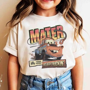 K&ouml;nnte beinhalten: Cremefarbenes T-Shirt mit einem Aufdruck von Hook aus dem Film Cars. Das Design zeigt den Namen "MATER" in gro&szlig;en, farbigen Buchstaben &uuml;ber einem Bild der Figur. Darunter steht der Text "JUST KEEP TRUCKIN'" und "TOWING SALVAGE".