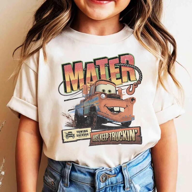 K&ouml;nnte beinhalten: Cremefarbenes T-Shirt mit einem Aufdruck von Hook aus dem Film Cars. Das Design zeigt den Namen "MATER" in gro&szlig;en, farbigen Buchstaben &uuml;ber einem Bild der Figur. Darunter steht der Text "JUST KEEP TRUCKIN'" und "TOWING SALVAGE".