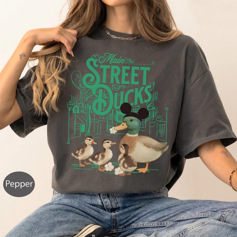 Op de afbeelding: Donkergrijs t-shirt met een groene afbeelding van "Main Street Ducks Co." met een eend met een Mickey Mouse-hoed en drie eendenkuikens. Het ontwerp is geplaatst tegen een groene straat.