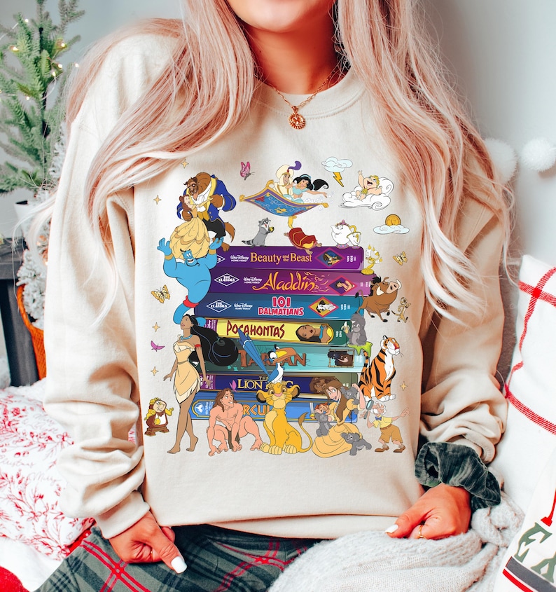 Puede incluir: Sudadera beige con una pila de libros coloridos con t&iacute;tulos y personajes de pel&iacute;culas de Disney. Los libros incluyen "La Bella y la Bestia", "Aladd&iacute;n", "101 D&aacute;lmatas", "Pocahontas" y "El Rey Le&oacute;n". Varios personajes de dibujos animados est&aacute;n representados.
