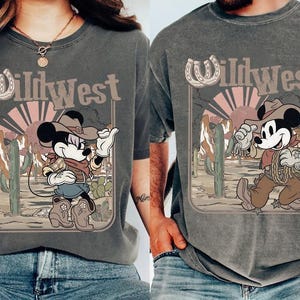 May include: Two gray t-shirts featuring a cartoon Mickey Mouse in a cowboy outfit with the text "Wild West". The design includes a horseshoe, cacti, and a desert sunset. The shirts are worn by two people.