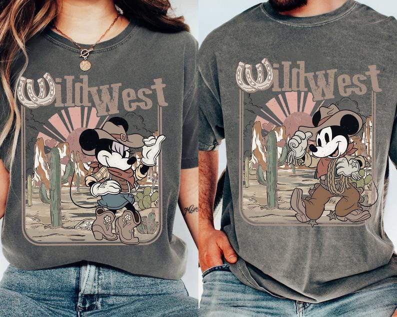 May include: Two gray t-shirts featuring a cartoon Mickey Mouse in a cowboy outfit with the text "Wild West". The design includes a horseshoe, cacti, and a desert sunset. The shirts are worn by two people.
