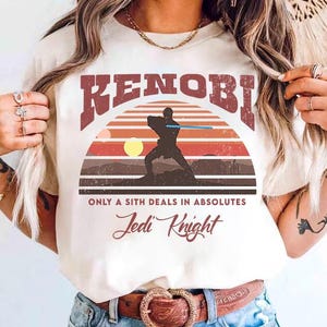 Op de afbeelding: Crèmekleurig T-shirt met retro-ontwerp. Het woord "KENOBI" in bordeauxrood boven een silhouet van een Jedi Knight met een lichtzwaard tegen een zonsondergang. De tekst luidt "ONLY A SITH DEALS IN ABSOLUTES" en "Jedi Knight".