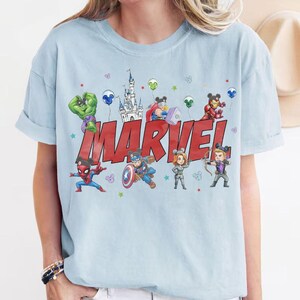 May include: Light blue t-shirt featuring the word "MARVEL" in red block letters, with cartoon illustrations of Marvel characters. The characters include Spider-Man, Captain America, and others. The shirt has rolled-up sleeves.