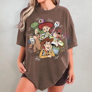 May include: A brown short-sleeve t-shirt featuring a graphic print of various animated characters including Woody, Jessie, Rex, Bullseye, and Buzz Lightyear. The design includes handwritten-style names and text such as Hi and Slinky Dog.