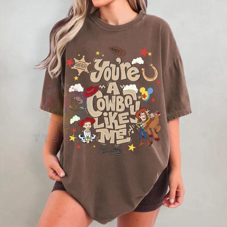 Pu&ograve; includere: Una t-shirt marrone a maniche corte con una stampa grafica che riporta il testo "You're a cowboy like me" e "Andy" su uno stivale. Il design include illustrazioni di Woody, Jessie e Bullseye, insieme a stelle, nuvole e un lazo come elementi grafici.