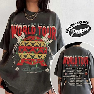 May include: A charcoal gray short-sleeve t-shirt featuring a graphic of a globe with hands making rock signs, wrapped in string lights. The text reads "Drink Around The World Tour" with a list of cities and countries including Mexico, Norway, and Orlando, FL.