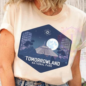 Puede incluir: Camiseta color crema con un dise&ntilde;o hexagonal azul marino. El dise&ntilde;o muestra un paisaje futurista con una gran luna, edificios y una estructura espacial. El texto dice "Tomorrowland National Park."