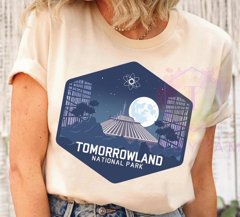 Puede incluir: Camiseta color crema con un dise&ntilde;o hexagonal azul marino. El dise&ntilde;o muestra un paisaje futurista con una gran luna, edificios y una estructura espacial. El texto dice "Tomorrowland National Park."