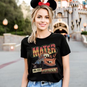 K&ouml;nnte beinhalten: Schwarzes T-Shirt mit einem Aufdruck von Hook aus dem Film Cars, mit den Worten "MATER" und "JUST KEEP TRUCKIN'". Das Design zeigt eine orange, blaue und braune Abschleppwagen-Illustration. Die Person tr&auml;gt ein Minnie-Maus-Ohren-Stirnband.