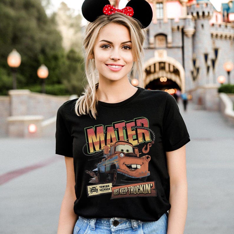 K&ouml;nnte beinhalten: Schwarzes T-Shirt mit einem Aufdruck von Hook aus dem Film Cars, mit den Worten "MATER" und "JUST KEEP TRUCKIN'". Das Design zeigt eine orange, blaue und braune Abschleppwagen-Illustration. Die Person tr&auml;gt ein Minnie-Maus-Ohren-Stirnband.