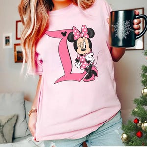 Peut inclure: Un t-shirt &agrave; manches courtes rose clair arborant un grand graphisme de la lettre D rose avec un personnage de souris de dessin anim&eacute; portant une robe et un n&oelig;ud &agrave; pois roses. Le personnage est plac&eacute; &agrave; c&ocirc;t&eacute; de la lettre, incluant une ic&ocirc;ne d'oreille.