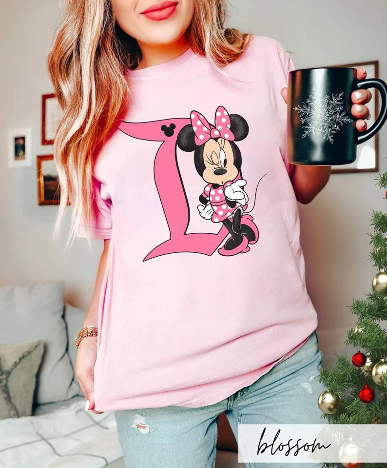 Peut inclure: Un t-shirt &agrave; manches courtes rose clair arborant un grand graphisme de la lettre D rose avec un personnage de souris de dessin anim&eacute; portant une robe et un n&oelig;ud &agrave; pois roses. Le personnage est plac&eacute; &agrave; c&ocirc;t&eacute; de la lettre, incluant une ic&ocirc;ne d'oreille.