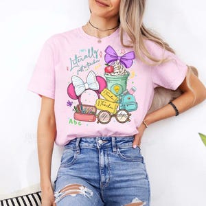 May include: Pink t-shirt with a colorful graphic design featuring a Minnie Mouse-inspired bow, a coffee cup with whipped cream, a backpack, and the text "Literally just a teacher". The design includes pencils, glasses, and apple.