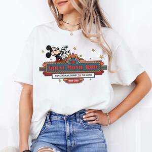 Puede incluir: Camiseta blanca con un gr&aacute;fico colorido de Mickey Mouse sosteniendo una claqueta de cine sobre el texto "The Great Movie Ride". El dise&ntilde;o incluye estrellas y el texto "A Spectacular Journey Through The Movies 1989-2017".