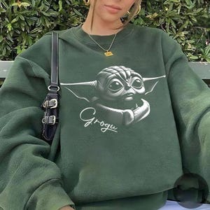 K&ouml;nnte beinhalten: Dunkelgr&uuml;nes Sweatshirt mit einem schwarz-wei&szlig;en Grogu-Motiv, dem Charakter aus The Mandalorian, und dem Namen "Grogu" darunter. Das Sweatshirt ist einfarbig und hat einen Rundhalsausschnitt.