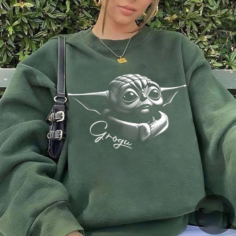 K&ouml;nnte beinhalten: Dunkelgr&uuml;nes Sweatshirt mit einem schwarz-wei&szlig;en Grogu-Motiv, dem Charakter aus The Mandalorian, und dem Namen "Grogu" darunter. Das Sweatshirt ist einfarbig und hat einen Rundhalsausschnitt.