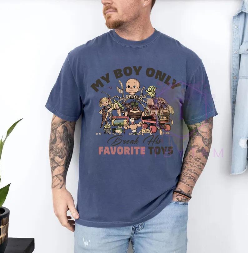 K&ouml;nnte beinhalten: Ein blaues kurz&auml;rmeliges T-Shirt mit einem Grafikdruck verschiedener Spielzeugfiguren und dem Text "MY BOY ONLY" oben sowie "Break His FAVORITE TOYS" unten in brauner und orangefarbener Schrift, mit Rundhalsausschnitt und lockerer Passform.