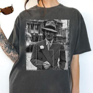 May include: Dark grey t-shirt with a black and white image of a man in a suit and hat. The image is framed by white lines and the letters "WALT". The shirt has a vintage, worn appearance.