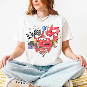 May include: White t-shirt featuring a red cartoon race car with the number 5 and the name "Jacob". The design includes checkered flags, balloons, and a birthday hat, celebrating a fifth birthday.
