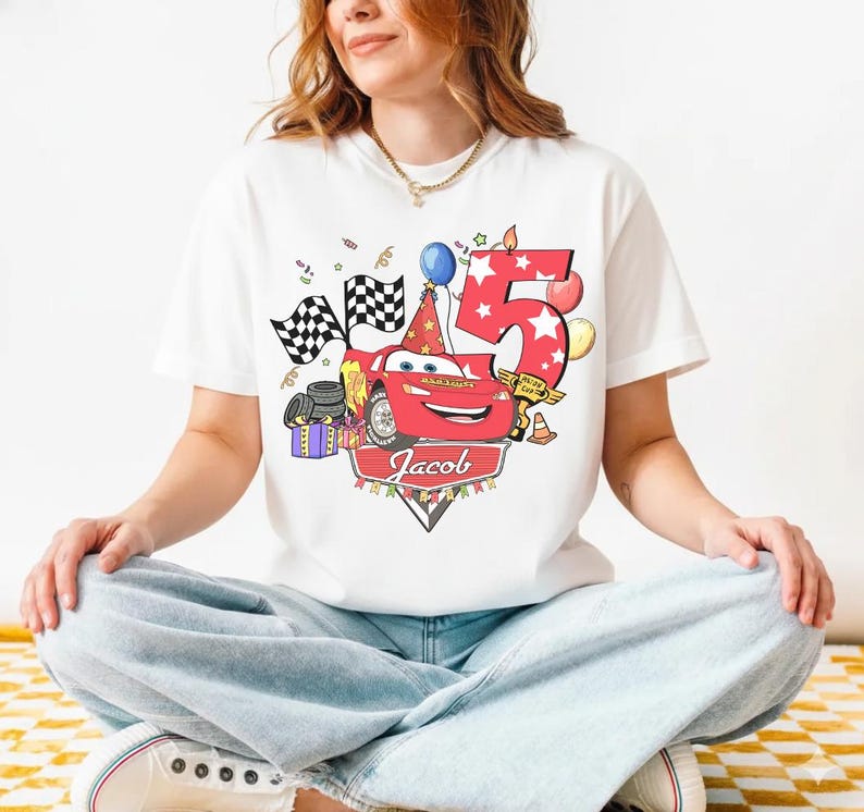 May include: White t-shirt featuring a red cartoon race car with the number 5 and the name "Jacob". The design includes checkered flags, balloons, and a birthday hat, celebrating a fifth birthday.