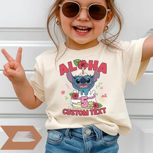 May include: Cream-colored t-shirt featuring a cartoon character with the text "Aloha" and "5". The graphic includes a cake and the words "Custom Text". The shirt is worn by a child.
