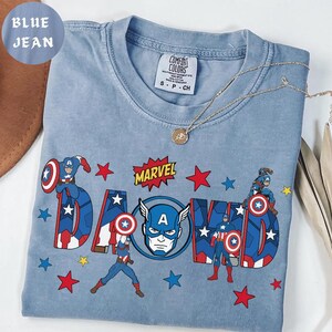 May include: A blue jean colored t-shirt featuring Captain America graphics and the word "DAD" in a bold, comic book style. The shirt includes the Marvel logo and red and blue star accents. A gold necklace is draped across the shirt.