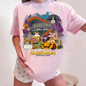 K&ouml;nnte beinhalten: Ein hellrosa T-Shirt mit kurzen &Auml;rmeln und einer bunten Grafik von Pixar Pier im California Adventure Park, die verschiedene Zeichentrickfiguren in Halloween-Kost&uuml;men zeigt, darunter ein K&uuml;rbis-Auto und Mickey-Mouse-Ohren.