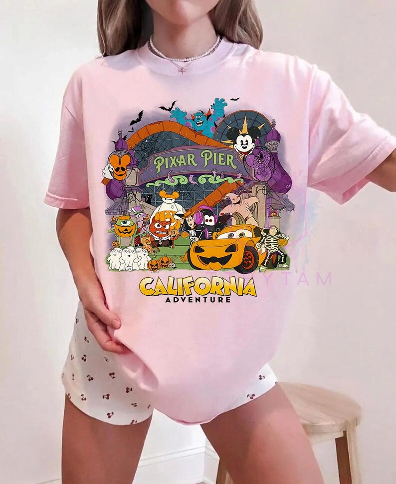 K&ouml;nnte beinhalten: Ein hellrosa T-Shirt mit kurzen &Auml;rmeln und einer bunten Grafik von Pixar Pier im California Adventure Park, die verschiedene Zeichentrickfiguren in Halloween-Kost&uuml;men zeigt, darunter ein K&uuml;rbis-Auto und Mickey-Mouse-Ohren.