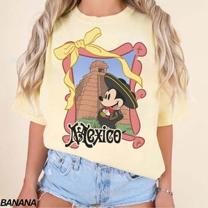 May include: Yellow t-shirt featuring a framed graphic of Mickey Mouse in a sombrero and traditional Mexican attire, with a pyramid in the background. The word "Mexico" is written in a decorative font below the image. A yellow bow is at the top of the frame.