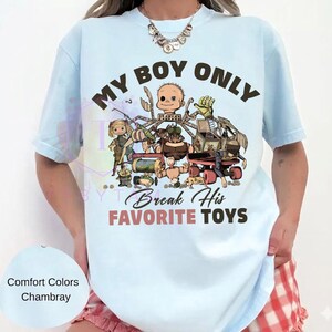 K&ouml;nnte beinhalten: Ein hellblaues T-Shirt mit einem Grafikdruck verschiedener Spielzeugfiguren und dem Text "MY BOY ONLY" in Dunkelbraun sowie "Break His FAVORITE TOYS" in Dunkelbraun und gedecktem Rot. Das Design zeigt unter anderem einen Puppenkopf und eine Spieluhr.