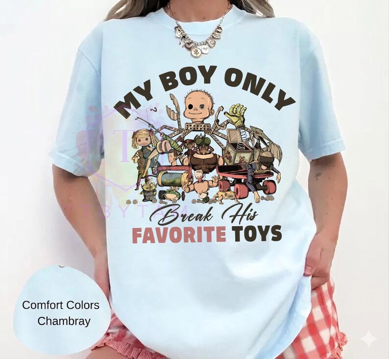 K&ouml;nnte beinhalten: Ein hellblaues T-Shirt mit einem Grafikdruck verschiedener Spielzeugfiguren und dem Text "MY BOY ONLY" in Dunkelbraun sowie "Break His FAVORITE TOYS" in Dunkelbraun und gedecktem Rot. Das Design zeigt unter anderem einen Puppenkopf und eine Spieluhr.
