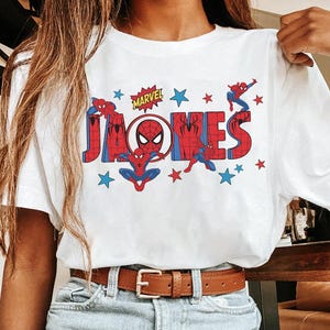 May include: White t-shirt featuring a colorful graphic of Spider-Man and the name "JONES" in red and blue block letters. The Marvel logo is above the name, with stars scattered around the design.