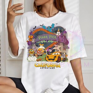 K&ouml;nnte beinhalten: Ein wei&szlig;es Kurzarm-T-Shirt mit einer bunten Grafik von Pixar Pier im California Adventure Park, die verschiedene Zeichentrickfiguren in Halloween-Kost&uuml;men zeigt, darunter ein k&uuml;rbisf&ouml;rmiges Auto, Geister und Mickey Mouse, mit dem Text Pixar Pier.