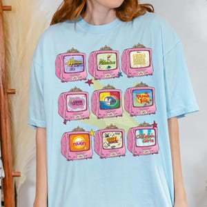 May include: Light blue t-shirt featuring a grid of nine pink retro televisions. Each TV displays a different show title, including "Hannah Montana," "Kim Possible," "High School Musical," "Lizzie McGuire," "Girl Meets World," "Phineas and Ferb," "Raven," "Camp Rock," and "Cheetah Girls."