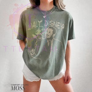 Może przedstawiać: Koszulka z kr&oacute;tkim rękawem w kolorze mchu z nadrukiem graficznym przedstawiającym Walta Disneya i Myszkę Miki. Napis "WALT DISNEY" jest nadrukowany jasną czcionką nad ilustracją. Koszulka ma okrągły dekolt i luźny kr&oacute;j.