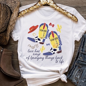 Pu&ograve; includere: Una t-shirt bianca a maniche corte con una stampa grafica di due stivali da cowboy gialli e blu con dettagli maculati, un cappello rosso, cactus e un fiocco. Il testo recita: "love has ways of bringing things back to life" in blu scuro.