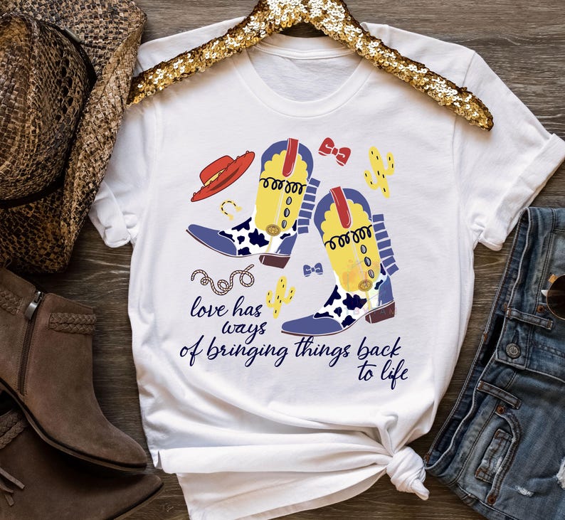 Pu&ograve; includere: Una t-shirt bianca a maniche corte con una stampa grafica di due stivali da cowboy gialli e blu con dettagli maculati, un cappello rosso, cactus e un fiocco. Il testo recita: "love has ways of bringing things back to life" in blu scuro.