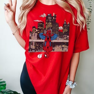 May include: A red t-shirt featuring a graphic of Spider-Man hanging upside down in front of a cityscape. The design includes a question mark speech bubble. The shirt is a vibrant red color.