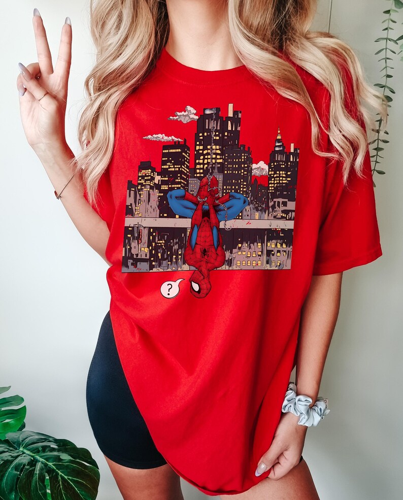 May include: A red t-shirt featuring a graphic of Spider-Man hanging upside down in front of a cityscape. The design includes a question mark speech bubble. The shirt is a vibrant red color.