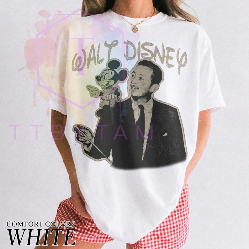 Może przedstawiać: Biały t-shirt z kr&oacute;tkim rękawem z grafiką w stylu vintage przedstawiającą Walta Disneya w ciemnym garniturze trzymającego batutę, z małą figurką Myszki Miki na ramieniu. Napis WALT DISNEY jest nadrukowany w postarzanym stylu.
