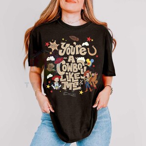 Pu&ograve; includere: Una t-shirt nera a maniche corte con una stampa grafica che riporta il testo "You're a cowboy like me" in un carattere color beige. Il design include illustrazioni di una stella da sceriffo, un ferro di cavallo, uno stivale e due personaggi.