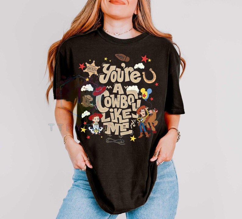 Pu&ograve; includere: Una t-shirt nera a maniche corte con una stampa grafica che riporta il testo "You're a cowboy like me" in un carattere color beige. Il design include illustrazioni di una stella da sceriffo, un ferro di cavallo, uno stivale e due personaggi.