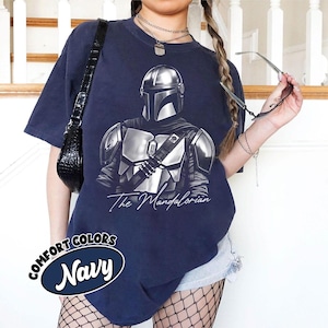 K&ouml;nnte beinhalten: Marineblaues T-Shirt mit einer detaillierten Illustration von The Mandalorian. Die Grafik ist in Grau- und Silbert&ouml;nen gehalten, mit dem Text "The Mandalorian" in einer Schriftart. Das Shirt hat auch den Text "Comfort Colors Navy" in einem kreisf&ouml;rmigen Design.