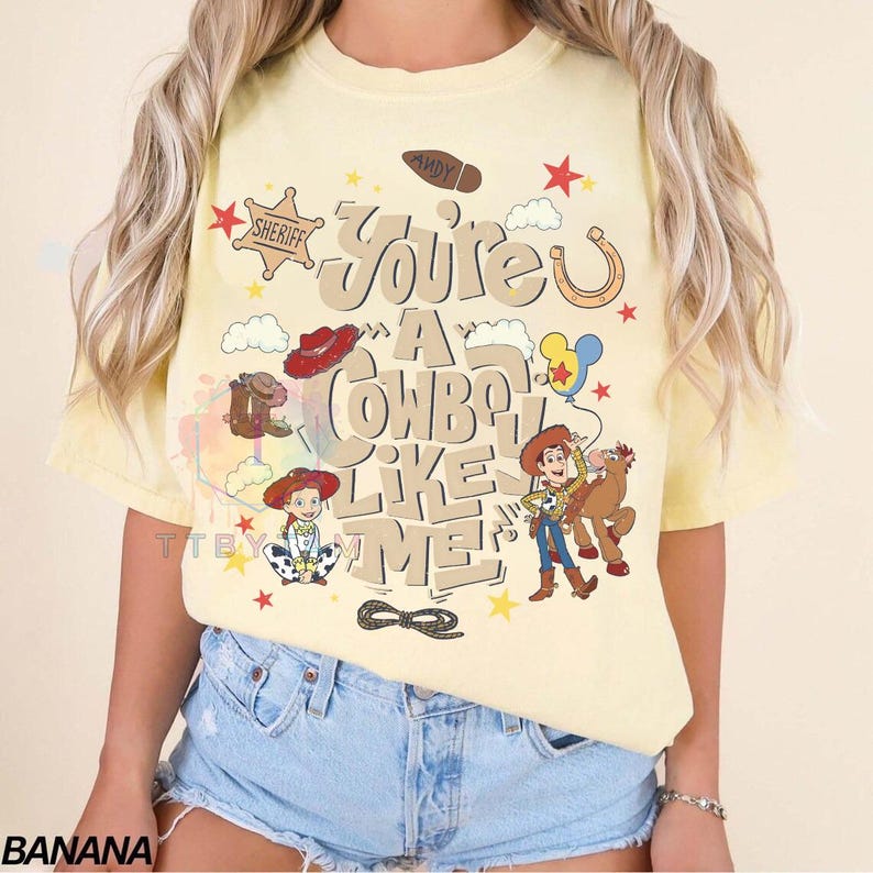 Pu&ograve; includere: Una t-shirt giallo pallido con una stampa grafica che riporta il testo "You're A Cowboy Like Me" e "Andy" sulla suola di uno stivale. Il design include illustrazioni di una cowgirl, un cowboy, un cavallo, una stella da sceriffo e stelle rosse.