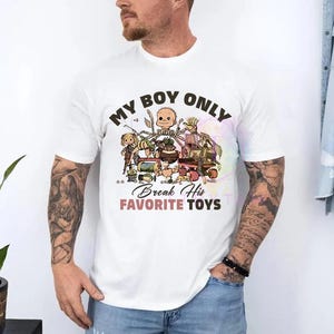 K&ouml;nnte beinhalten: Ein wei&szlig;es kurz&auml;rmeliges T-Shirt mit einem Grafikdruck verschiedener Spielzeugfiguren. Der Text lautet oben MY BOY ONLY und unten Break His FAVORITE TOYS in schwarzer und rosa Schrift. Das Design enth&auml;lt in der Mitte einen Puppenkopf.