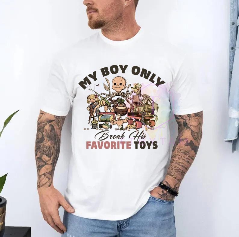 K&ouml;nnte beinhalten: Ein wei&szlig;es kurz&auml;rmeliges T-Shirt mit einem Grafikdruck verschiedener Spielzeugfiguren. Der Text lautet oben MY BOY ONLY und unten Break His FAVORITE TOYS in schwarzer und rosa Schrift. Das Design enth&auml;lt in der Mitte einen Puppenkopf.